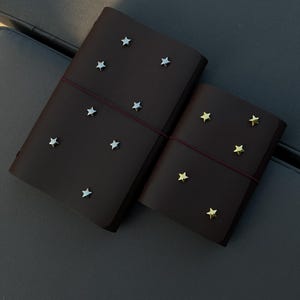 Könnte beinhalten: Zwei dunkelbraune Lederjournale mit Sternverzierungen. Ein Journal hat silberne Sterne, das andere goldene Sterne. Beide werden mit einem roten Gummiband verschlossen.