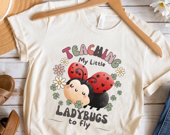 May include: A beige t-shirt with a graphic of a ladybug with the text "Teaching My Little Ladybugs to Fly".