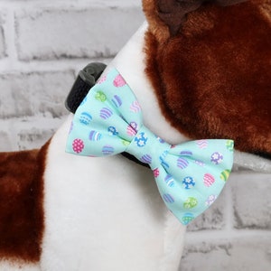 May include: A plush dog toy with brown and white fur, wearing a light blue bow tie decorated with colorful Easter egg designs. The dog is positioned in front of a white brick wall.