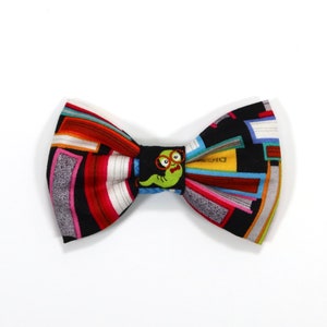 May include: A black bow tie with a colorful book pattern and a green worm with glasses. The worm is in the center of the bow tie and is surrounded by books.