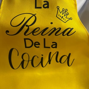 May include: A bright yellow apron with black text and a crown graphic. The text reads "La Reina De La Cocina". The apron has a simple design with a neck strap and waist ties.