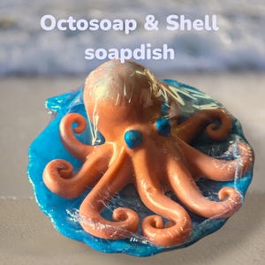May include: An orange octopus soap sits on a blue seashell-shaped soap dish. The soap dish is wrapped in clear plastic. The text "Octosoap & Shell soapdish" is at the top of the image.