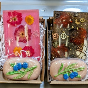 May include: Two gift boxes with clear resin trays containing pressed flowers and a bar of soap with a floral design. The first box has a pink tray with pressed flowers and a bar of soap with a green leaf and blue berries. The second box has a dark brown tray with gold flecks and a bar of soap with a green leaf and blue berries.