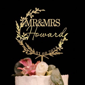 May include: Gold wedding cake topper with a floral wreath design. The topper says "MR & MRS Howard" and "01.08.2024".