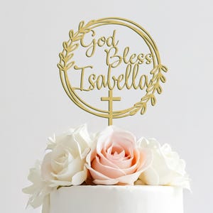 May include: A three-tiered white cake decorated with pink and white roses. A gold cake topper reads "God Bless Isabella" within a wreath and cross design. The cake is likely for a special occasion.