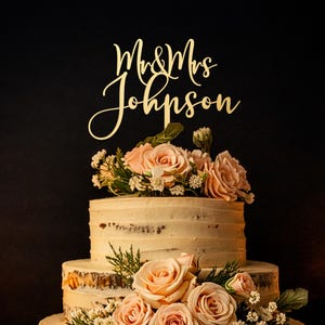 May include: A gold-colored cake topper with the text Mr & Mrs Johnson in a cursive script font, positioned atop a three-tiered cake decorated with light peach-colored roses, small white flowers, and green foliage on a wooden cake stand.