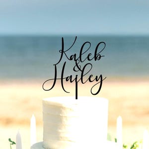 May include: A black cake topper featuring the names Kaleb & Hailey in a cursive script font. The topper is positioned on top of a white frosted cake. The design consists of the name Kaleb above the name Hailey with an ampersand in between.