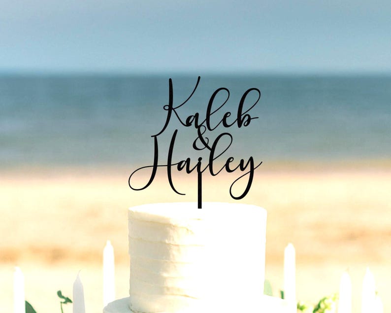 May include: A black cake topper featuring the names Kaleb & Hailey in a cursive script font. The topper is positioned on top of a white frosted cake. The design consists of the name Kaleb above the name Hailey with an ampersand in between.