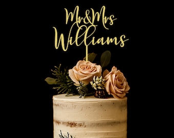 Puede incluir: Un adorno para pastel de color dorado con el texto Mr & Mrs Williams en una fuente cursiva. El adorno está insertado en un pastel redondo de color claro, decorado con dos rosas de color melocotón claro y follaje verde en la parte superior.