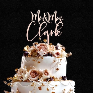 May include: A rose gold cake topper featuring the text Mr & Mrs Clark in a cursive script font, positioned atop a two-tiered white cake decorated with cream and light pink roses, small dark berries, and delicate gold-colored leafy vine accents.