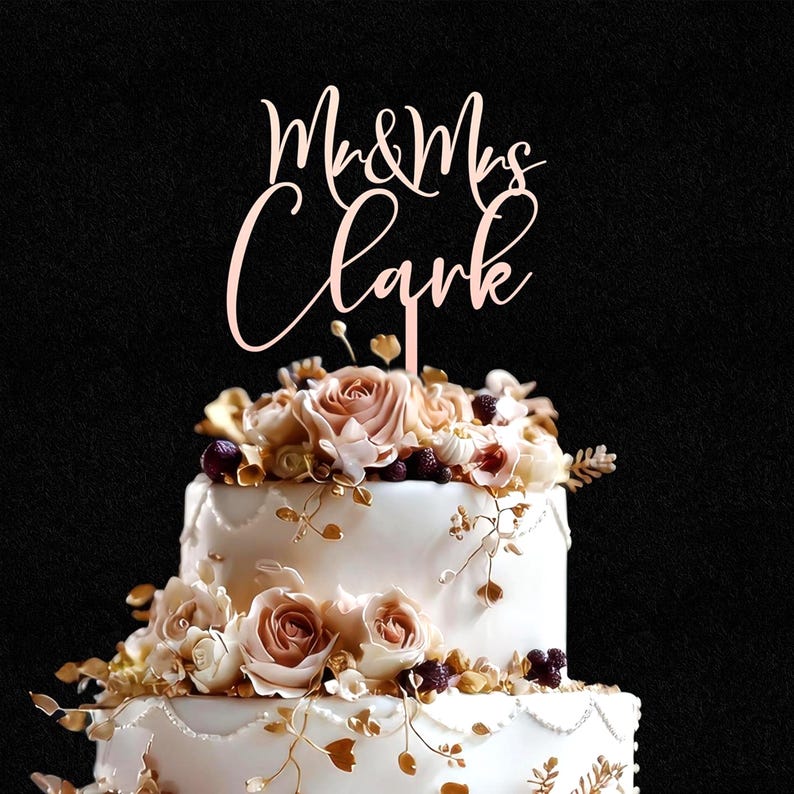 May include: A rose gold cake topper featuring the text Mr & Mrs Clark in a cursive script font, positioned atop a two-tiered white cake decorated with cream and light pink roses, small dark berries, and delicate gold-colored leafy vine accents.