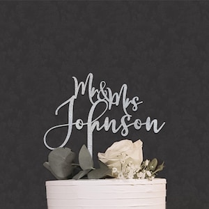 May include: A silver glitter cake topper featuring the text Mr & Mrs Johnson in a cursive script font. The topper is positioned on top of a white frosted cake decorated with a white rose and green leaves against a dark gray background.