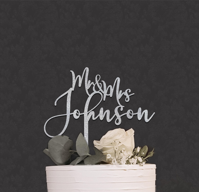 May include: A silver glitter cake topper featuring the text Mr & Mrs Johnson in a cursive script font. The topper is positioned on top of a white frosted cake decorated with a white rose and green leaves against a dark gray background.