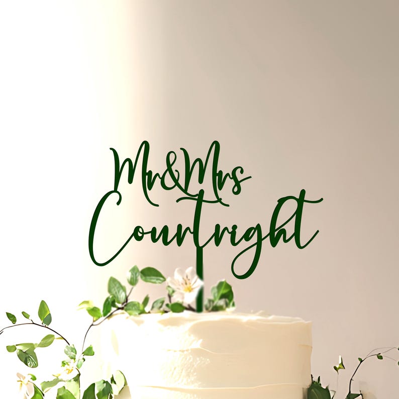 May include: A dark green cake topper featuring the text "Mr & Mrs Courtright" in a cursive script font. The topper is mounted on a thin vertical stake and is positioned atop a white frosted cake decorated with small green leaves and white flowers.