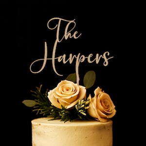 May include: A cake topper featuring the text The Harpers in a cursive script font, positioned above two light beige roses with green leaves, all resting on top of a smooth, light-colored frosted cake against a solid black background.
