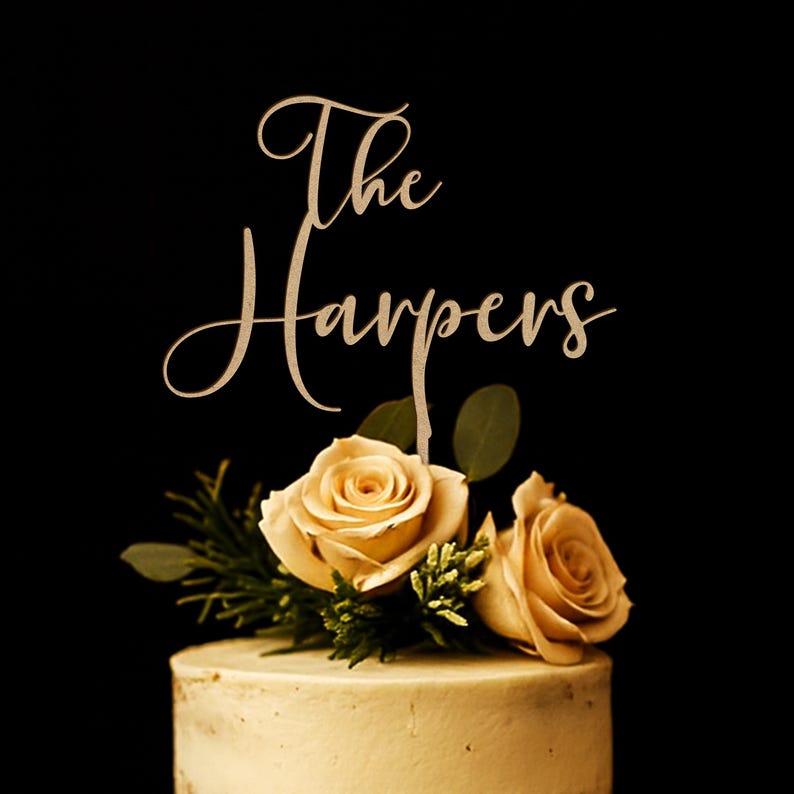 May include: A cake topper featuring the text The Harpers in a cursive script font, positioned above two light beige roses with green leaves, all resting on top of a smooth, light-colored frosted cake against a solid black background.