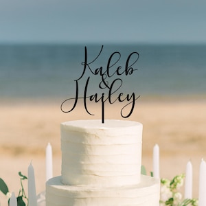 May include: A black cake topper featuring the names Kaleb & Hailey in a cursive script font. The topper is positioned on top of a white, multi-tiered cake with a textured, horizontal-lined frosting finish, creating a clear and legible display.