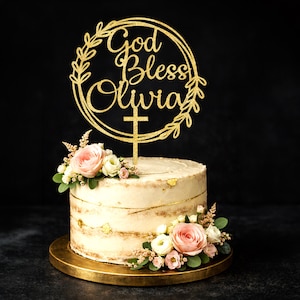 May include: A gold glitter cake topper featuring the text God Bless Olivia inside a circular frame with leaf accents and a cross at the bottom. The topper is placed on a round, frosted cake decorated with pink and white roses and green leaves.