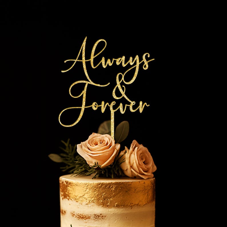 May include: A gold glitter cake topper featuring the cursive text Always & Forever, positioned atop a cake decorated with two light pink roses and green foliage. The topper has a thin vertical stake extending downward into the cake surface.