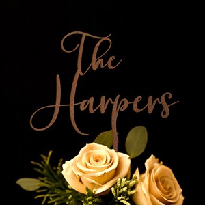 May include: A copper-colored metal cake topper featuring the text The Harpers in a cursive script font, positioned above two light peach-colored roses with green leaves and small sprigs of greenery against a solid black background.