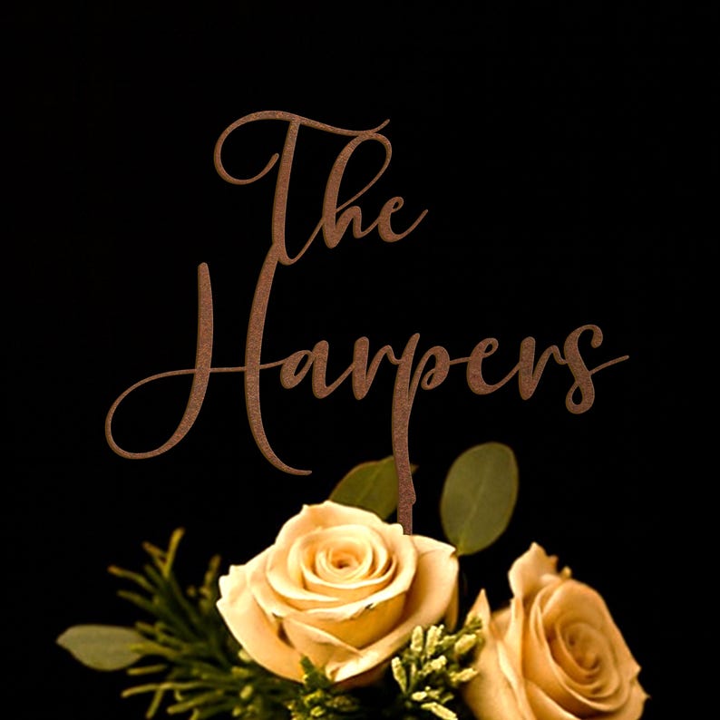 May include: A copper-colored metal cake topper featuring the text The Harpers in a cursive script font, positioned above two light peach-colored roses with green leaves and small sprigs of greenery against a solid black background.