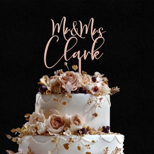 May include: A rose gold cake topper featuring the text Mr & Mrs Clark in a cursive script font, positioned atop a two-tiered white cake decorated with cream-colored roses, small dark berries, and delicate gold-toned leafy vine accents.
