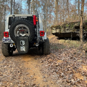 May include: A white Jeep Wrangler with a spare tire mounted on the back, parked on a dirt trail in a wooded area. The vehicle has black accents, red taillights, and a license plate. A black bag with white text hangs from the spare tire.