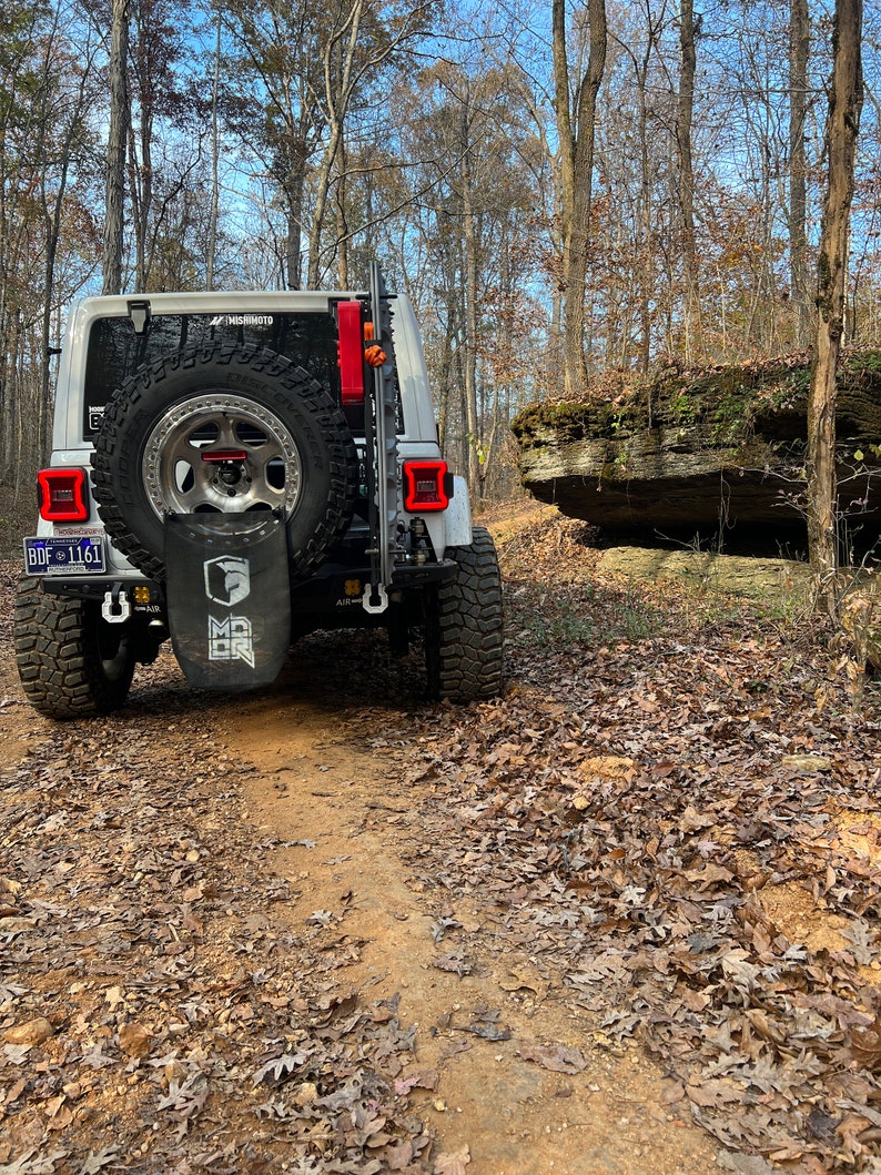 May include: A white Jeep Wrangler with a spare tire mounted on the back, parked on a dirt trail in a wooded area. The vehicle has black accents, red taillights, and a license plate. A black bag with white text hangs from the spare tire.