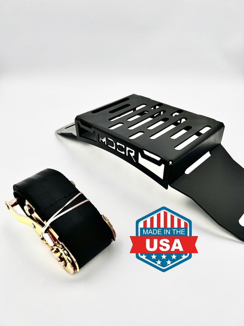May include: Black metal bracket with a slotted design and the word "MDCR" printed on it. A black strap with a ratchet mechanism and a "Made in the USA" emblem are also in the image.
