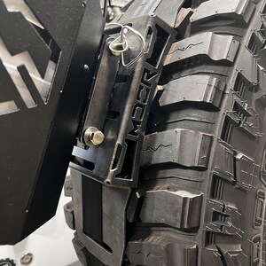 May include: Close-up of a black metal tire carrier mounted on a large off-road tire. The carrier has a geometric design with the word "AMBER" cut out. A silver pin secures a component. The tire has deep treads.