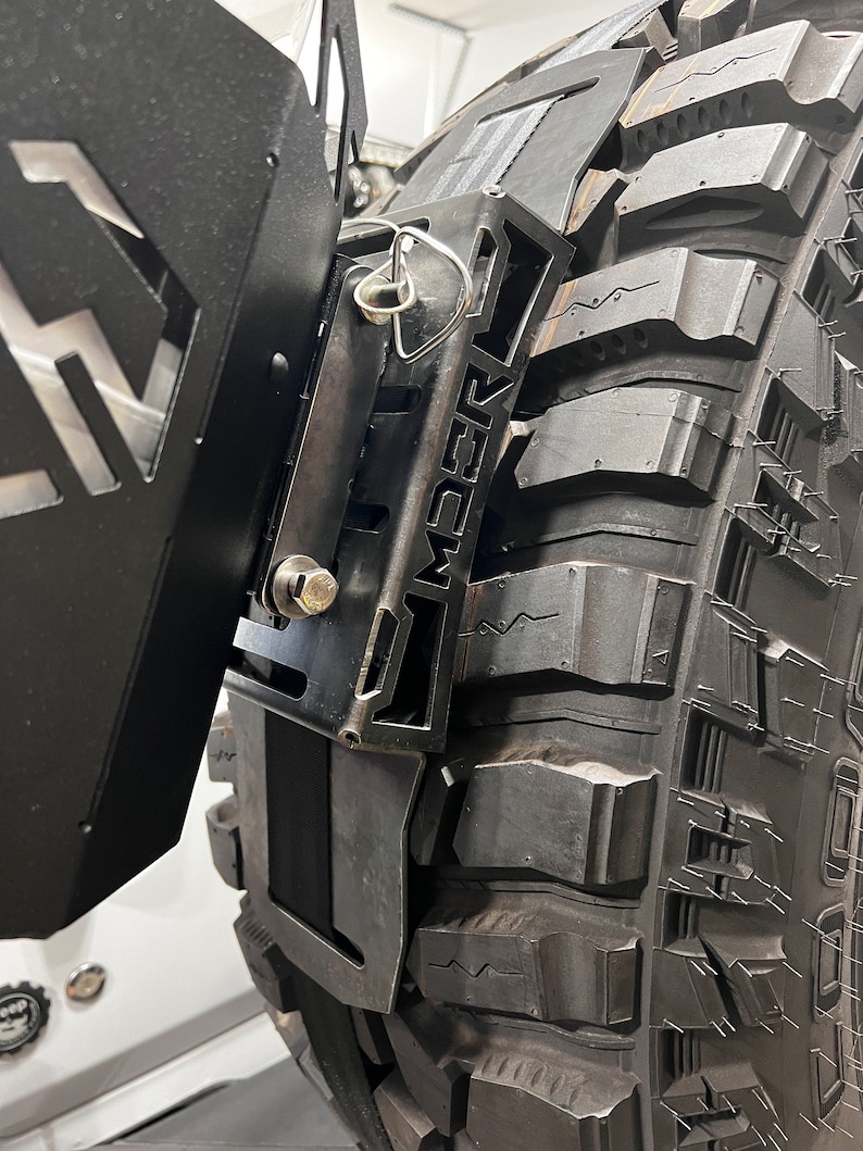 May include: Close-up of a black metal tire carrier mounted on a large off-road tire. The carrier has a geometric design with the word "AMBER" cut out. A silver pin secures a component. The tire has deep treads.