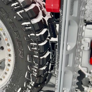May include: Close-up of a black off-road tire with the word "REAPER" and other text visible. The tire is covered in snow. A gray traction board is next to the tire, and a red object is visible in the background.