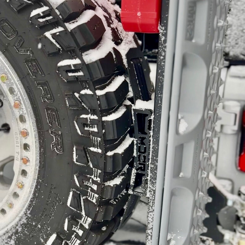 May include: Close-up of a black off-road tire with the word "REAPER" and other text visible. The tire is covered in snow. A gray traction board is next to the tire, and a red object is visible in the background.