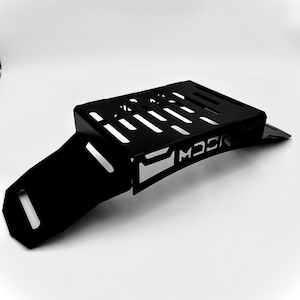 May include: A black metal motorcycle part with a rectangular top featuring several parallel cut-out slots. The part has a curved, angled extension and the word "MOCK" is cut out on the side.