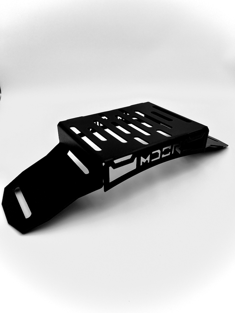 May include: A black metal motorcycle part with a rectangular top featuring several parallel cut-out slots. The part has a curved, angled extension and the word "MOCK" is cut out on the side.