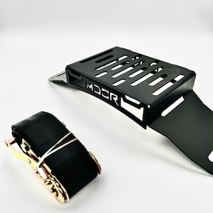 May include: Black metal bracket with a slotted top and the word "MDCR" printed on the side. A black ratchet strap with gold-colored hardware is next to the bracket. The items are on a white background.