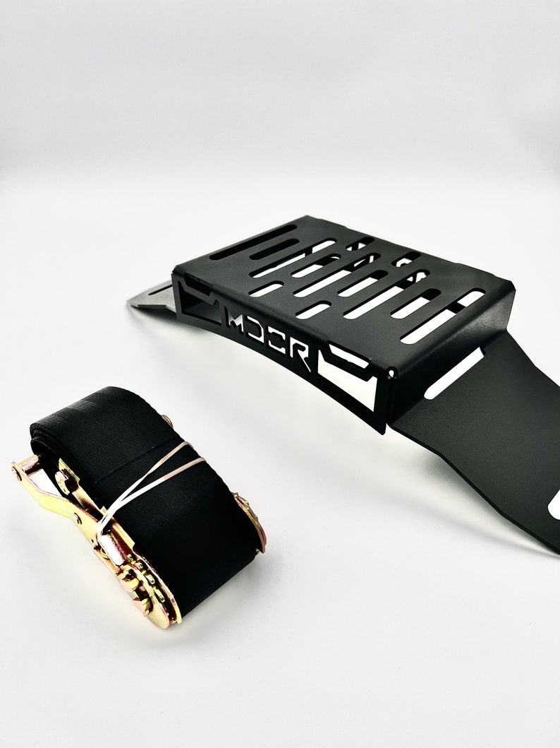 May include: Black metal bracket with a slotted top and the word "MDCR" printed on the side. A black ratchet strap with gold-colored hardware is next to the bracket. The items are on a white background.