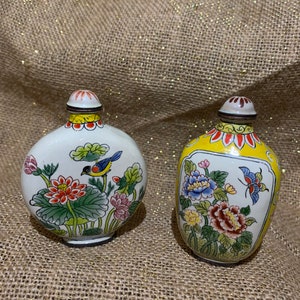 Pode incluir: Duas pequenas garrafas redondas de porcelana com intrincados desenhos florais e de pássaros. A garrafa à esquerda é branca com um pássaro azul e flores rosa e vermelhas. A garrafa à direita é amarela com uma borboleta e flores azuis, rosas e amarelas.