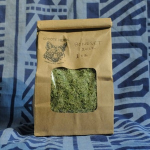 以下が含まれることがあります： 「Boneset 2022 1 oz」とラベルされた乾燥ハーブが入った茶色の紙袋。透明な窓から中の緑色のハーブが見えます。「Coyote Herbs」と「Snoqualmie Valley」のロゴが入っています。