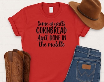 May include: A red t-shirt with the text "Some of y'alls CORNBREAD Ain't DONE IN the middle" is displayed with a brown cowboy hat, blue jeans, and brown cowboy boots. The words "NEW ARRIVAL" and "SHOP NOW" are also visible.