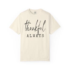 May include: Cream-colored t-shirt with the words "thankful ALWAYS" printed in black. The word "thankful" is in a cursive font, while "ALWAYS" is in a block font. The shirt has short sleeves and a crew neck.