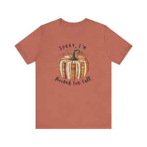 Peut inclure: Un t-shirt à manches courtes de couleur rouille chiné avec un motif de citrouille formée par des livres ouverts. Le texte au-dessus de la citrouille indique SORRY, I'M et le texte en dessous indique BOOKED FOR FALL dans une police sombre.