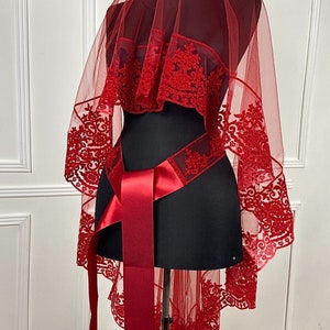 Puede incluir: Un velo de novia rojo con un delicado patrón de encaje y una cinta de satén a juego. El velo está hecho de tul y tiene un borde festoneado.