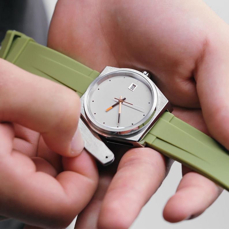May include: A silver watch with a white face and a green strap. The watch has a date window at the 3 o'clock position.