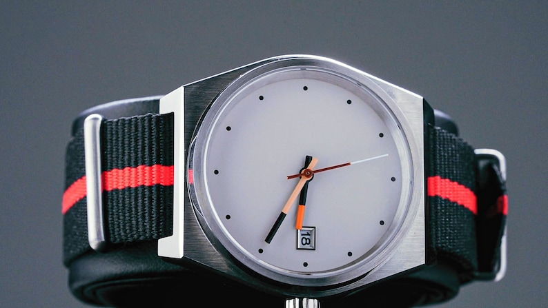 May include: A silver wristwatch with a white face and black and orange hands. The watch has a black and red striped nylon strap.