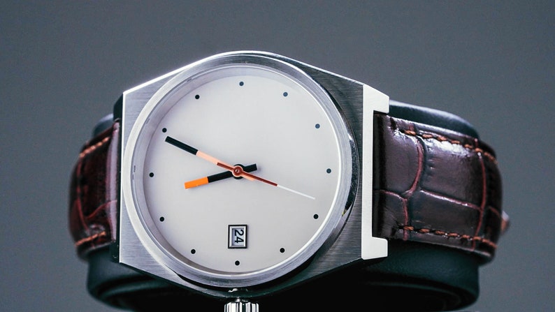 May include: A silver wristwatch with a white face and orange and black hands. The watch has a brown leather strap with a crocodile pattern.