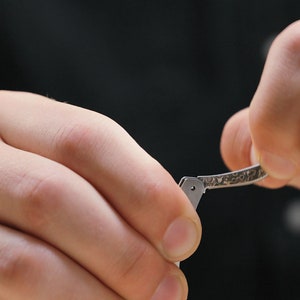 May include: A person's hands are holding a silver folding knife with a textured handle. The knife is being opened.