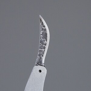 May include: A close-up of a silver pocket knife with a patterned blade. The blade is curved and has a sharp point.