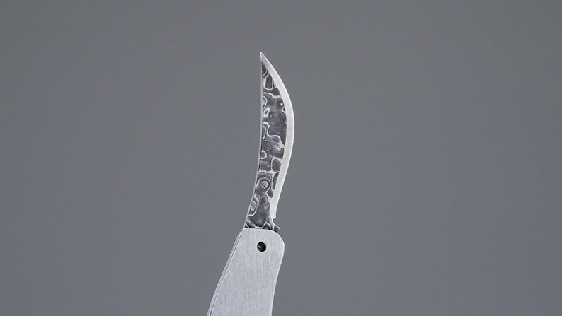 May include: A close-up of a silver pocket knife with a patterned blade. The blade is curved and has a sharp point.
