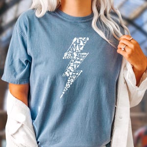May include: A blue t-shirt featuring a white lightning bolt design filled with various small illustrations. The person is wearing denim shorts and a white jacket. The background is a train station with a clock.